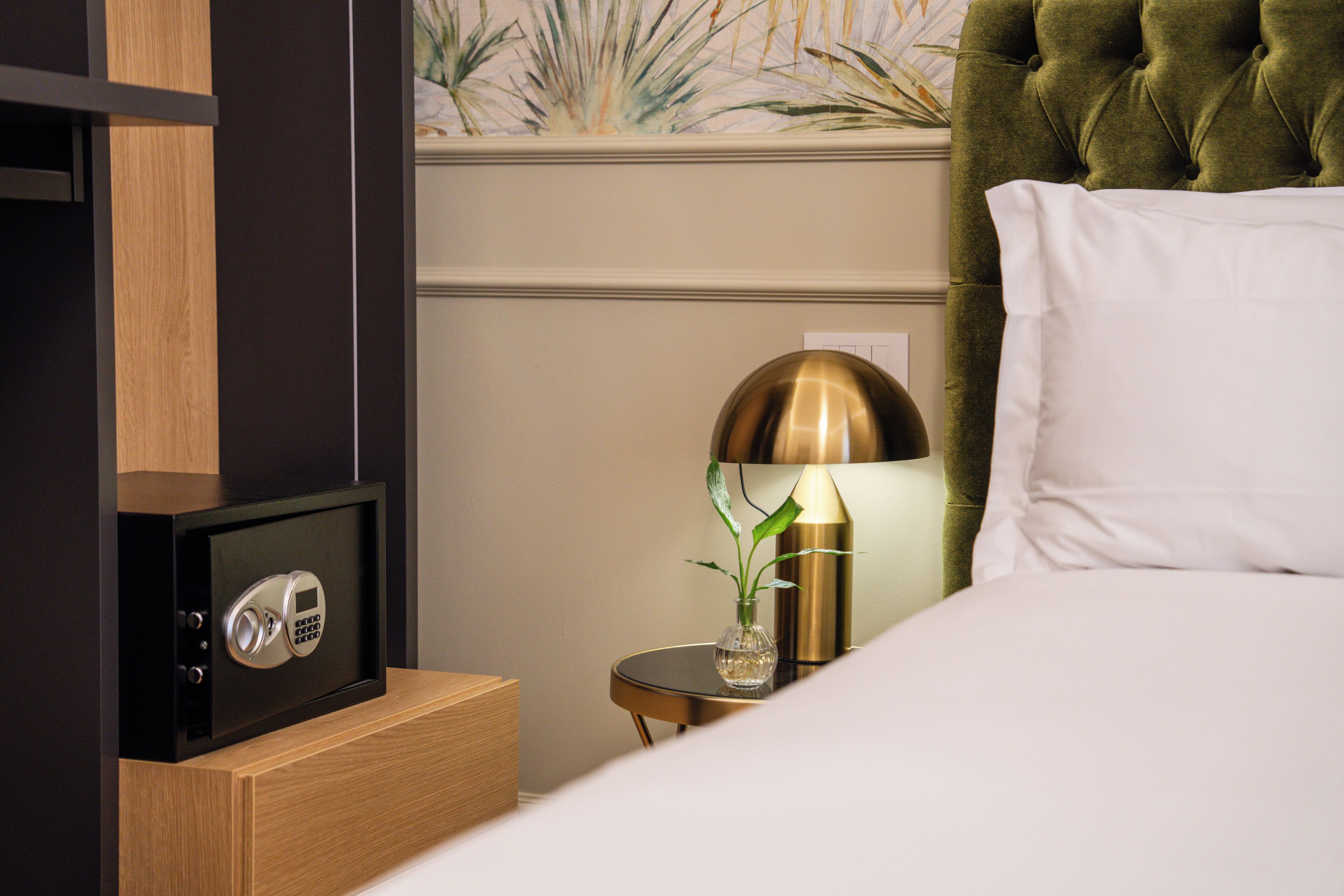 A hotel room with a bed, a safe, a small round table with a lamp, and a decorative plant.