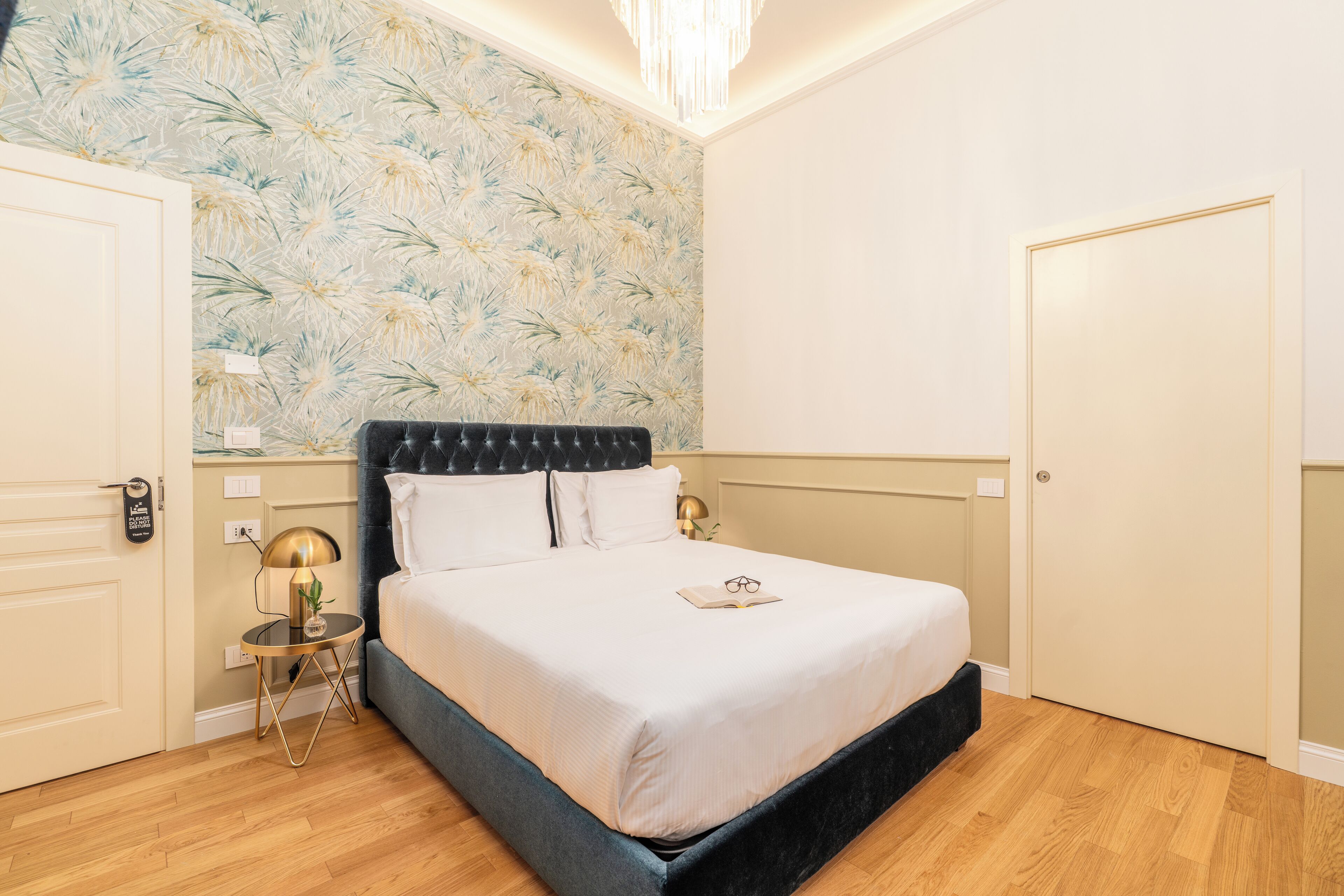 A neatly arranged bedroom with a bed, bedside lamps, and a floral wallpaper.