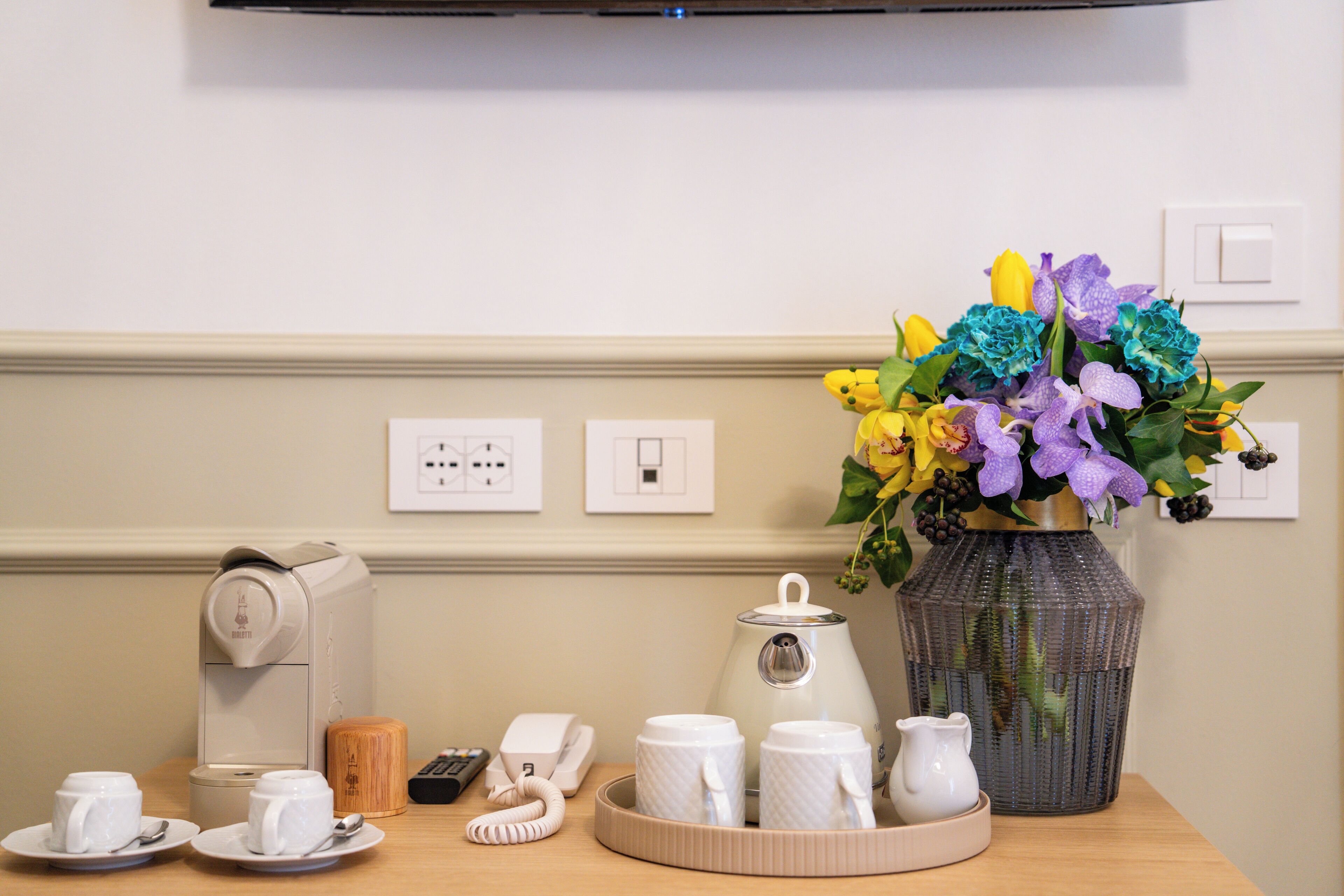 Una mesa ordenada con cafetera, teléfono, tazas, jarra y un florero con flores.