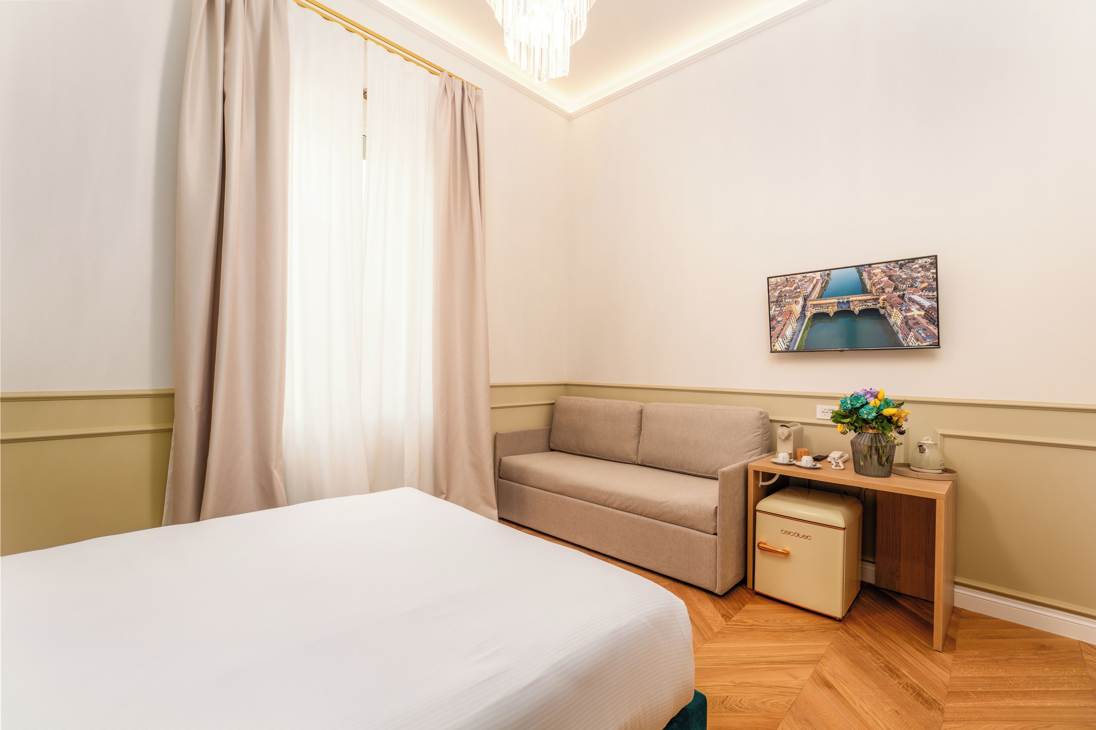 A hotel room with a bed, a sofa, a small table with a vase of flowers, and a painting on the wall.