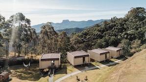 A modern resort with three cabins, each with a flat roof and a small porch. The cabins are surrounded by a well-maintained lawn and a hammock is visible. The resort is set against a backdrop of dense forest and rolling hills.