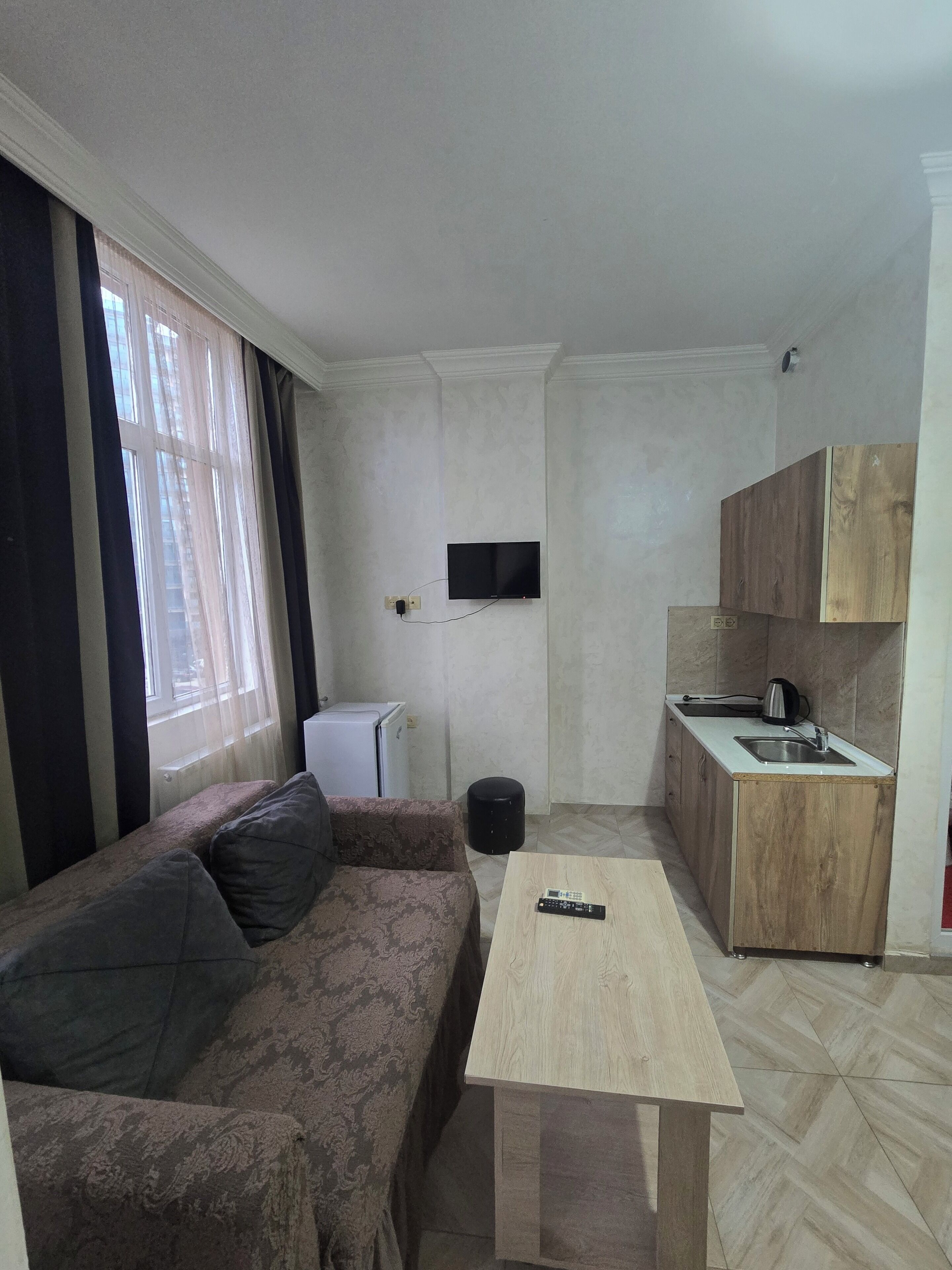 A compact living space with a sofa, a small table, a kitchen area with a sink and cabinets, a television mounted on the wall, and a window with curtains.