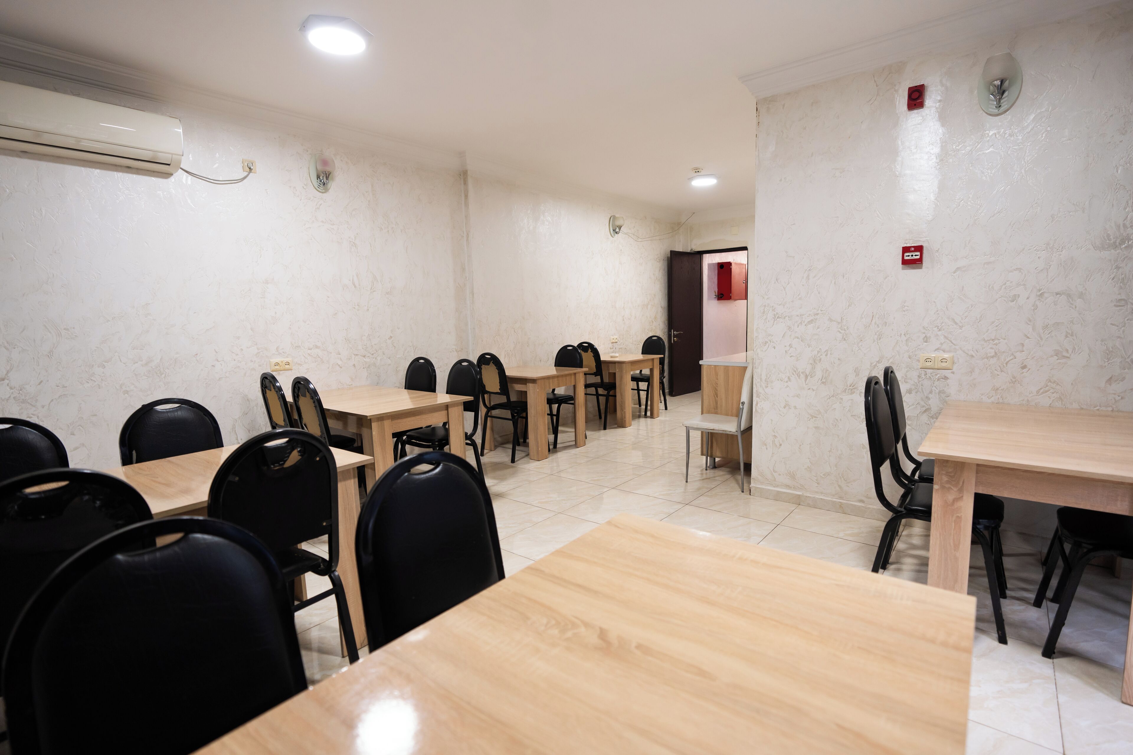 A conference room with multiple tables and chairs, air conditioning, and a door with a red emergency exit sign.