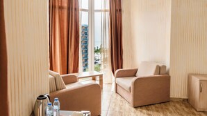 A hotel room with a sofa, a small table, and a cabinet. There is a window with curtains and a view of buildings outside. - Batumi Inn (Batumi)