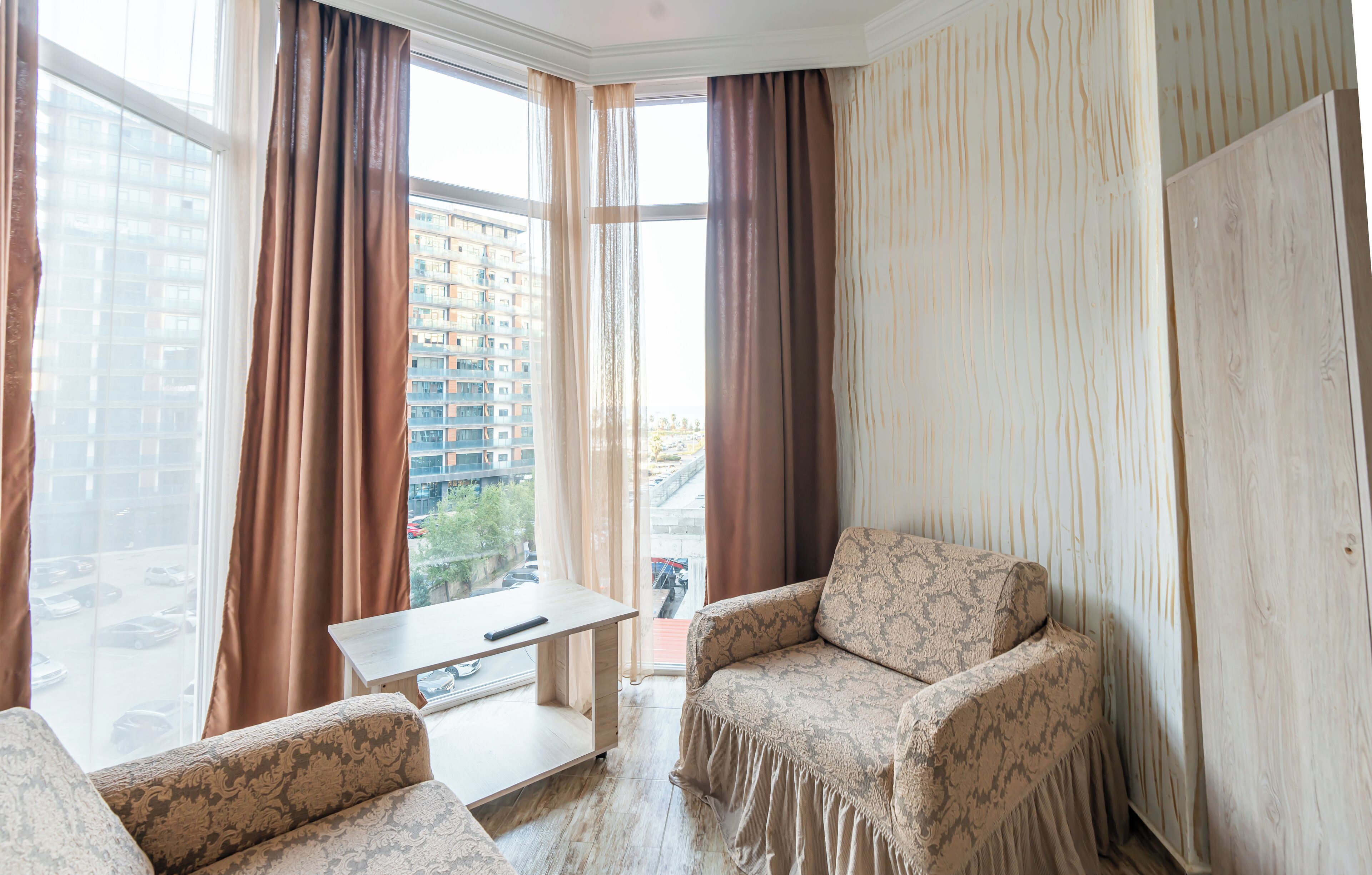 A corner room with a sofa, a small table, and a view of a cityscape through large windows with curtains.