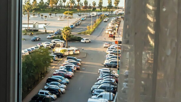 A view from a high-rise window overlooking a parking lot with cars, palm trees, and a body of water. - Batumi Inn (Batumi)