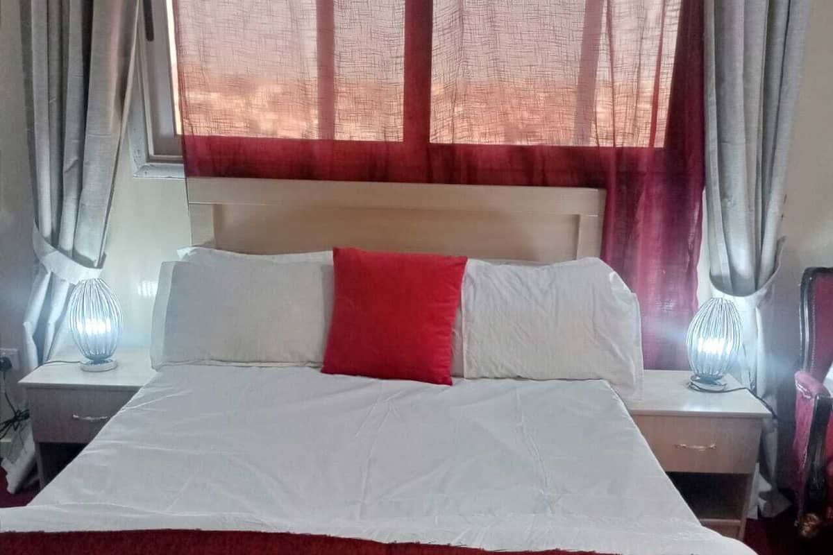 Een bed met wit en rood beddengoed, twee bedlampen en een raam met rode gordijnen.