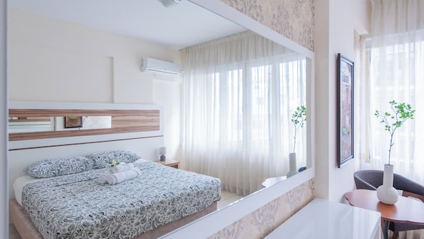 Une chambre à coucher moderne avec un grand lit, une table de chevet, un miroir et une fenêtre ornée de rideaux légers.