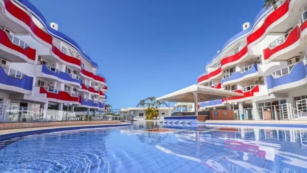 En swimmingpool med fliseterrasse, omgivet af høje bygninger med altaner.