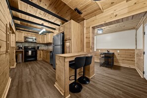 Una cocina de madera con una isla central, taburetes negros tipo barra, un refrigerador y un microondas.