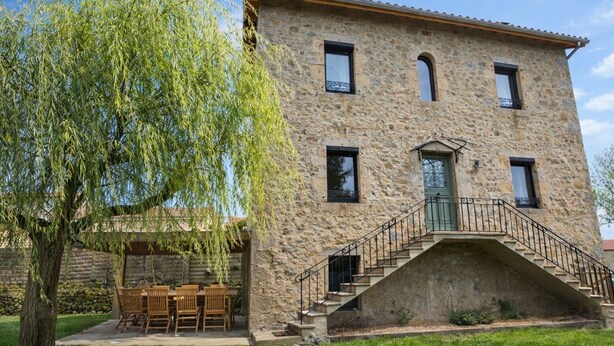 Un edificio de piedra con una escalera que conduce a una puerta verde, rodeado de césped y un árbol.