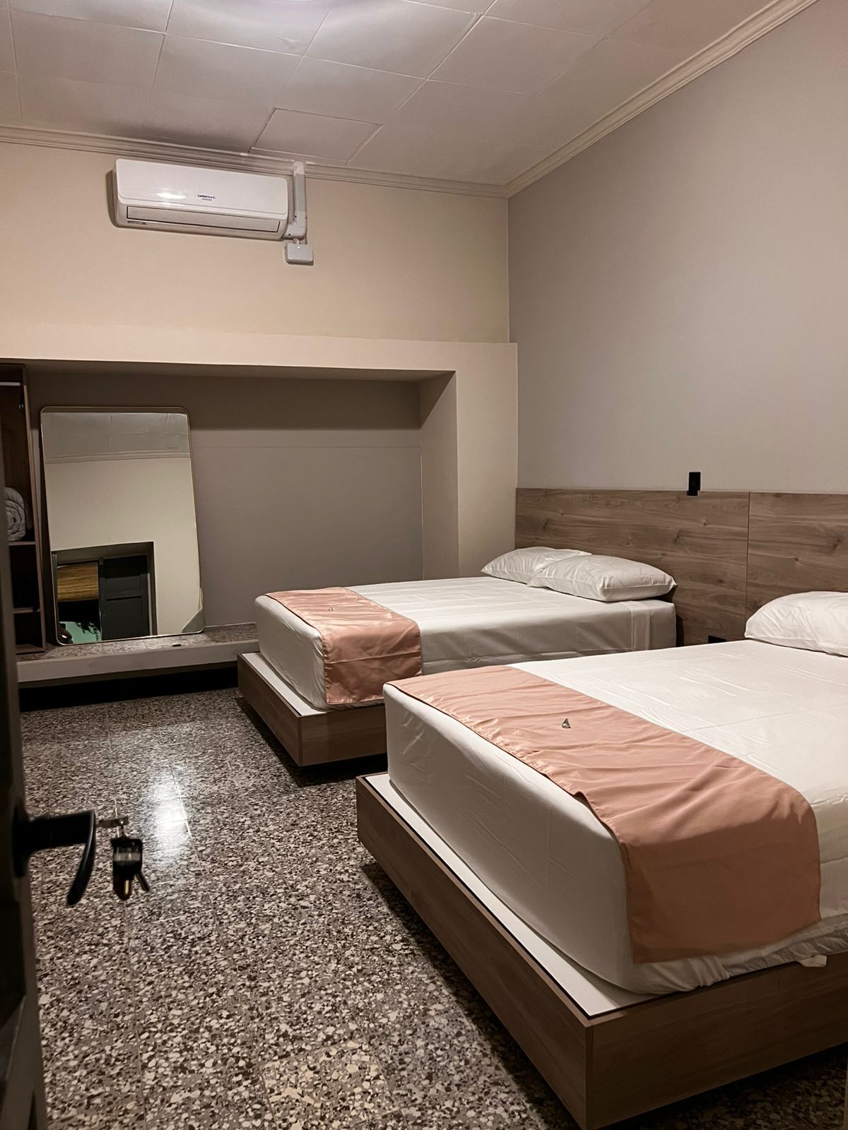 A hotel room with two beds, a wooden headboard, a mirror, and a ceiling-mounted air conditioning unit.