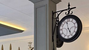 Reloj de pared con marco negro y esfera blanca, colgado de un pilar gris en una habitación con estantes que contienen objetos decorativos y un espejo.