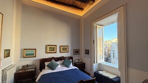 Een slaapkamer met een houten bed, een nachtkastje, een dressoir, een raam met uitzicht op de stad en lijstjes met foto's aan de muur.