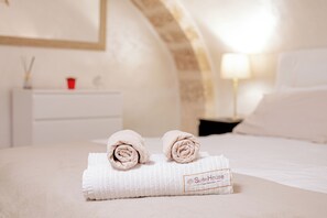 A neatly made bed with a white towel and a rolled towel on top. There is a nightstand with a lamp and a red cup. The room has a white color scheme and a framed picture on the wall. - SuiteHouse78 - Elegant, centrally located apartment with a private garden, near Bari's Old Town (Bari)
