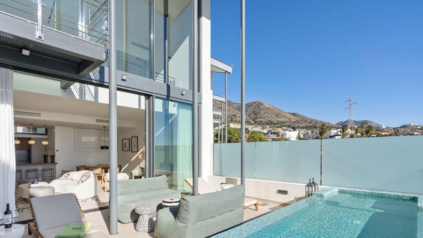 Ein moderner Poolbereich im Freien mit Glasgeländer, Essbereich mit Sofa und Tisch sowie Blick auf eine Hügelkette.