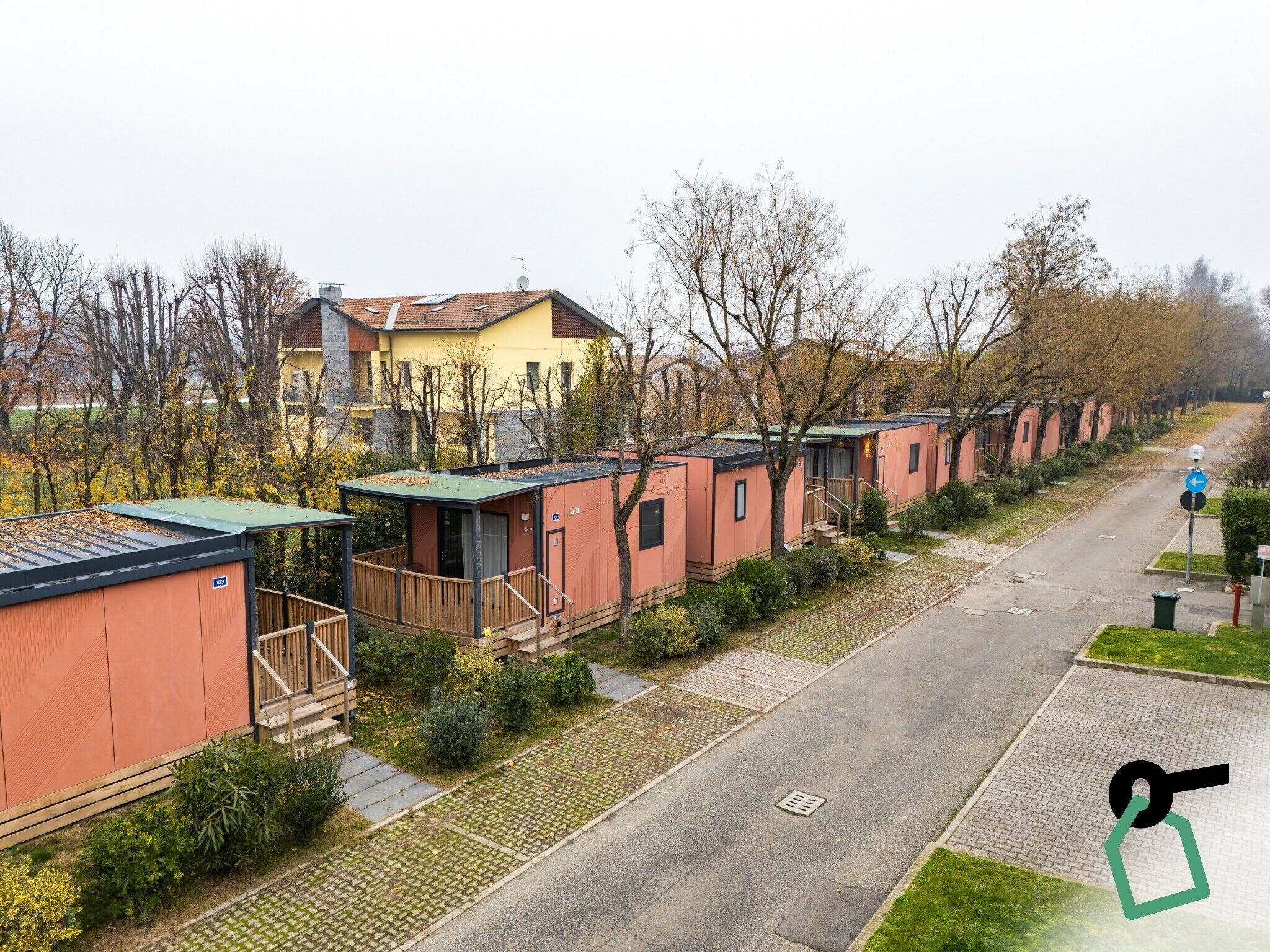 A row of small, single-story mobile homes with individual porches, a paved road, and a residential area with trees and a yellow house in the background.