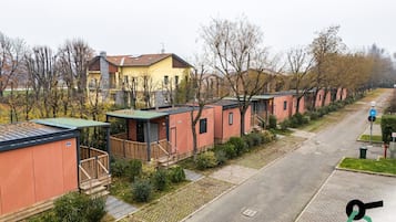 A row of small, single-story mobile homes with individual porches, a paved road, and a residential area with trees and a yellow house in the background.