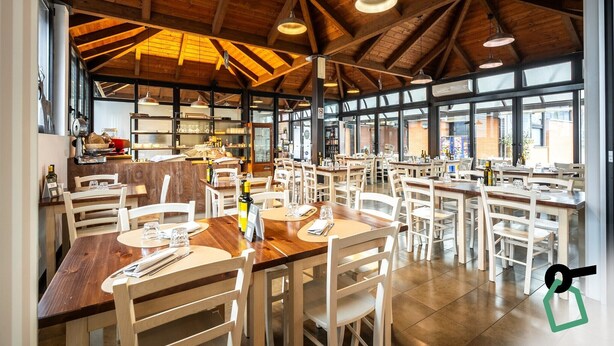 A spacious dining area with wooden tables and chairs, large windows, and a high ceiling with exposed beams. - Hotiday Bologna Arena (Bologna)