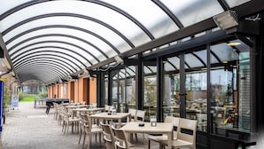 An outdoor cafe with tables and chairs, a grill, and a covered walkway with glass walls. - Hotiday Bologna Arena (Bologna)