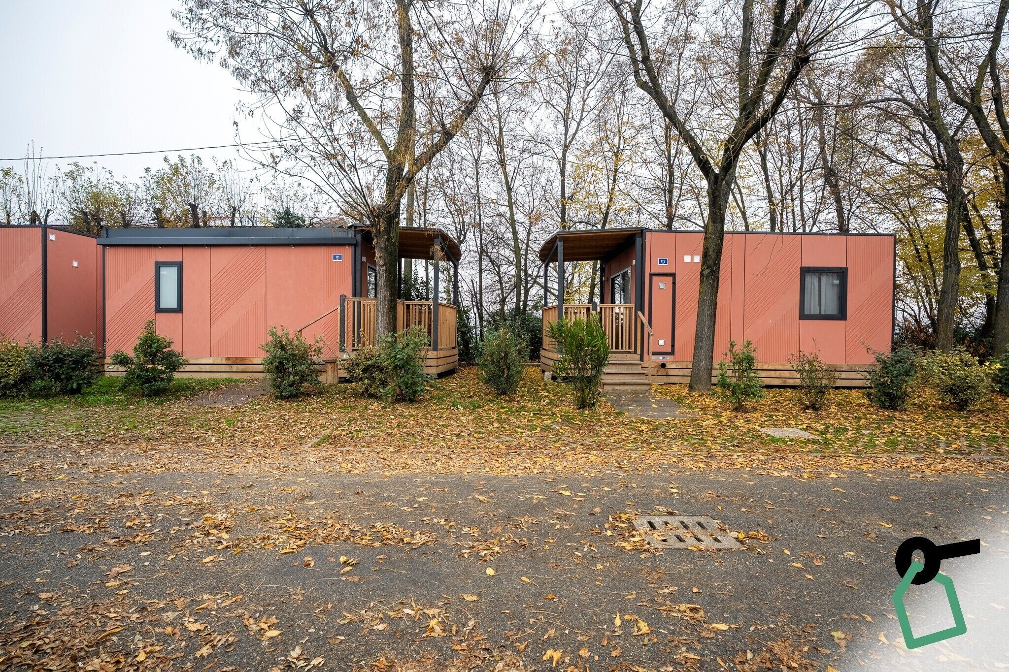 Twee moderne, compacte hutten met balkons, omringd door bomen en een parkeerplaats met herfstbladeren.