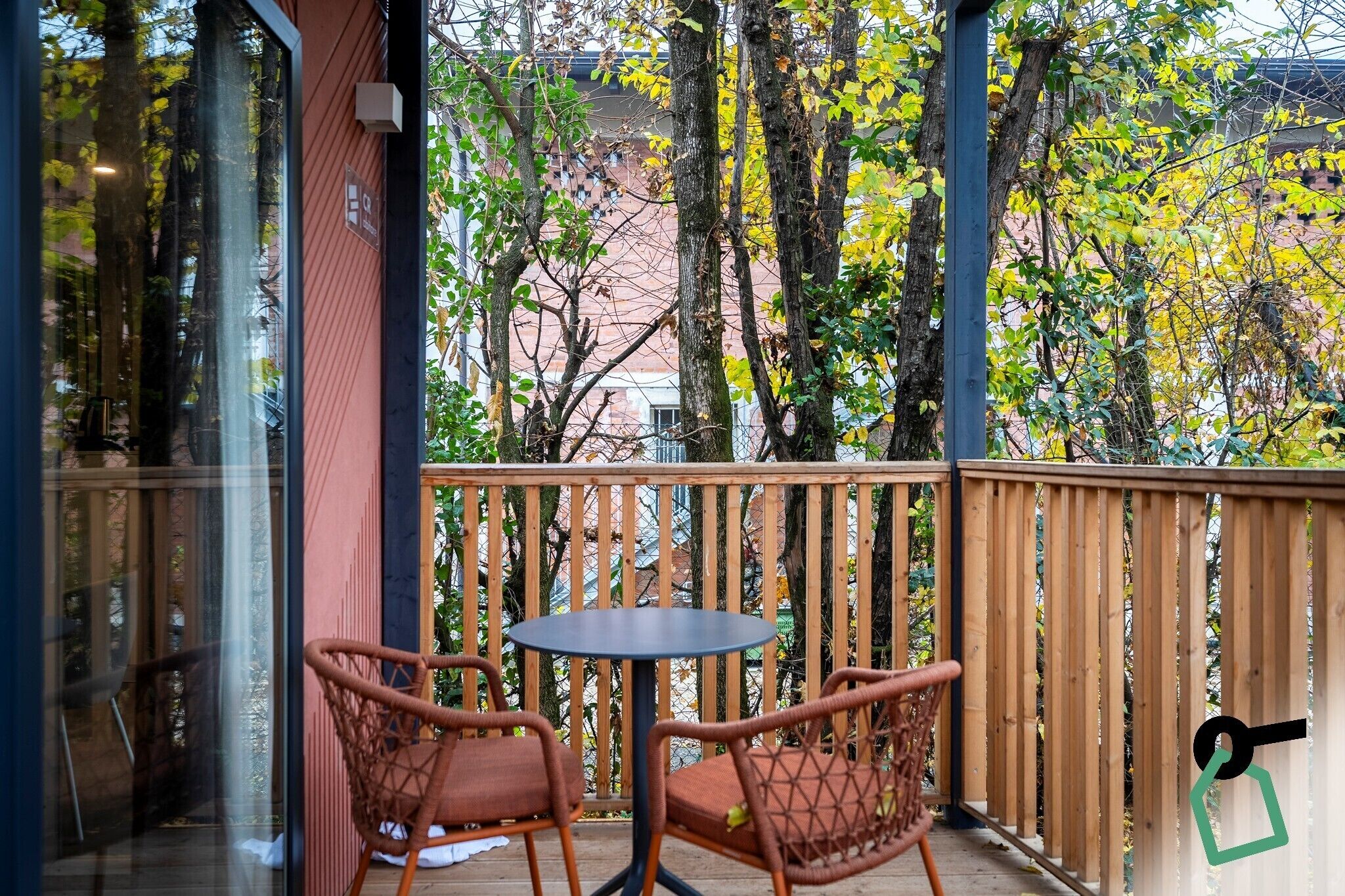 Un balcone con due sedie in vimini e un tavolino rotondo, circondato da ringhiere in legno e alberi con foglie gialle.