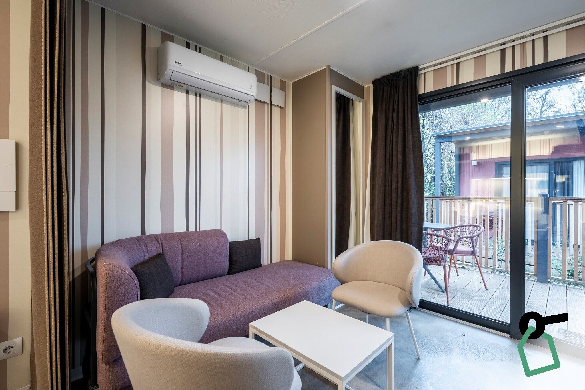 Een moderne woonkamer met een bank, twee fauteuils, een salontafel en een schuifdeur die uitkomt op een buitenruimte met uitzicht op een gebouw en bomen.
