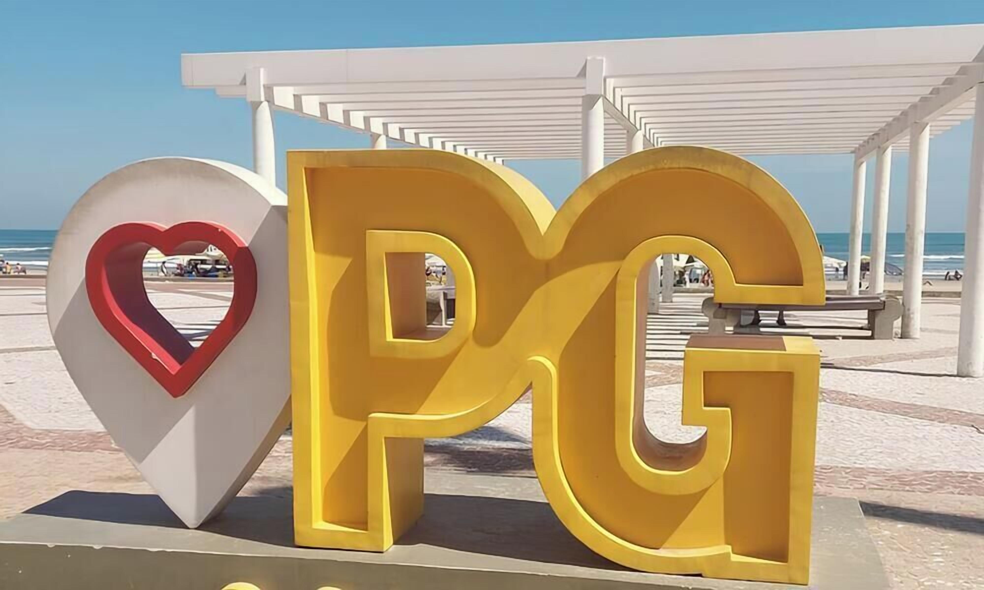 A large 'PG' sign with a heart-shaped cutout in the middle, located in a coastal area with a beach and a white pergola structure in the background.