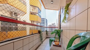 Ein schmaler Balkon mit Fliesenwänden, einem grünen Kunststoffstuhl, einem kleinen Tisch mit Flaschen und Snacks und Blick auf Gebäude durch ein Drahtgeflecht.