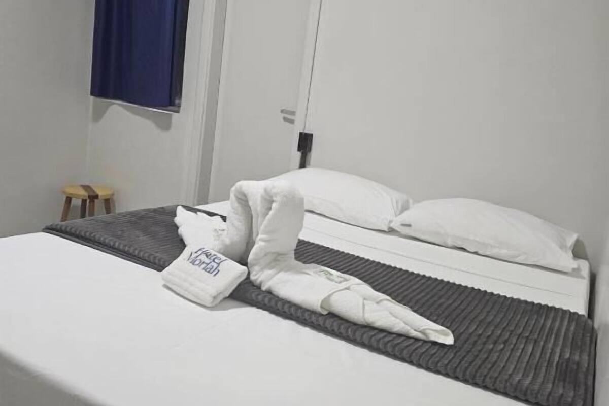 A hotel room with a bed, a small stool, a blue curtain, and an air conditioning unit.