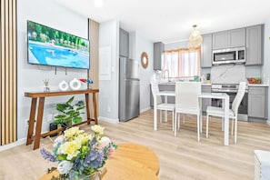 A modern kitchen with grey cabinets, a stainless steel refrigerator, a microwave, and a dining area with white chairs and a wooden table. - Honolulu Central 1 Bd 1 Ba (Honolulu)
