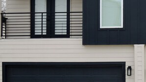 Een modern huis met twee verdiepingen, een zwarte garage deur, een klein raam en een lichtgekleurde gevel.