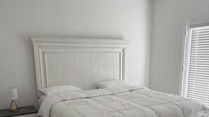 Een wit bed met een houten hoofdeinde, een nachtkastje met een lamp en een raam met jaloezieën.