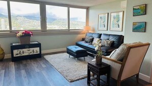 A living room with a sofa, a chair, a small table, and a rug. There are framed pictures on the wall and a window with a view of a cityscape. - Island Colony Waikiki High-Rise 1BR with Split AC & Full Kitchen (Honolulu)