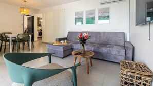 A modern living room with a grey sofa, a round wooden coffee table, and a green armchair. There is a dining area with a table and chairs, and a kitchen in the background. - 972L1 - T4 "Taïno" facing the sea (Le Vauclin)