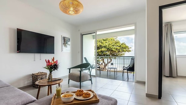 A modern living room with a flat-screen TV, a dining table with breakfast items, a sofa, and a balcony with outdoor furniture. - 972L1 - T4 "Taïno" facing the sea (Le Vauclin)
