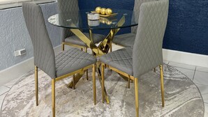Un espace repas comprenant une table en verre, quatre chaises grises à revêtement rembourré et un pied de table doré.