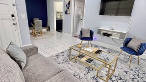 Un salon moderne avec un canapé gris, une table basse et deux fauteuils bleus. Un téléviseur est fixé au mur, et on aperçoit un coin cuisine au fond de la pièce.