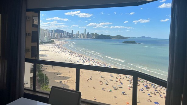 Vista a la playa desde una habitación con un ventanal grande, mostrando una playa de arena, el océano y el horizonte de una ciudad al fondo.