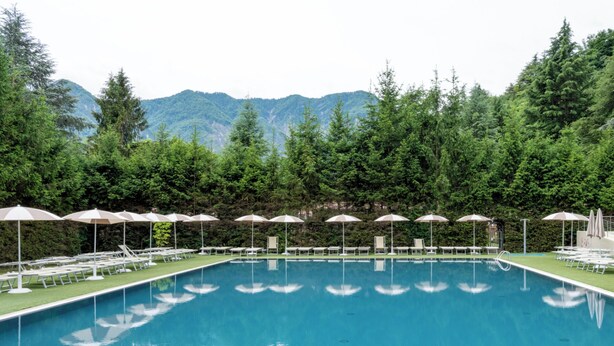 A swimming pool with lounge chairs and umbrellas, surrounded by trees and mountains. - Hotiday Lago di Levico (Levico Terme)