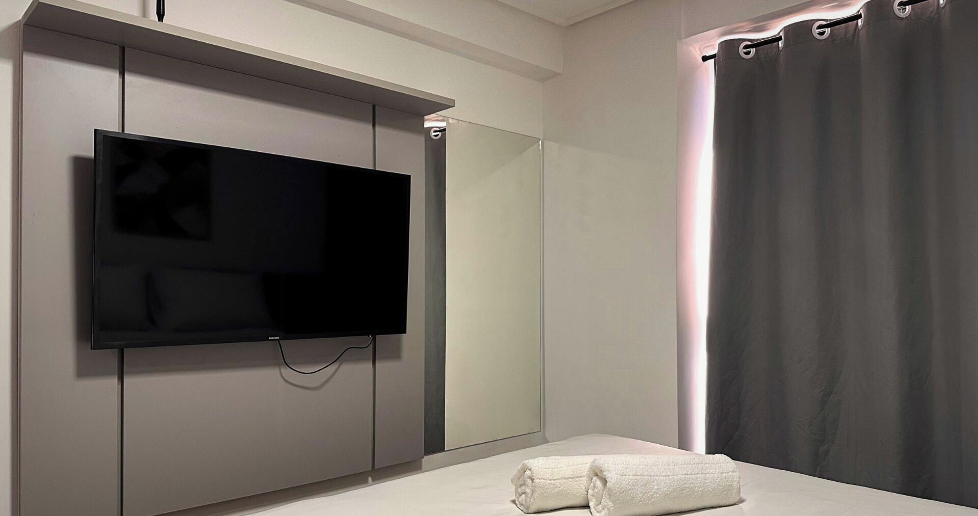 A modern bathroom with a flat-screen TV mounted on the wall, a towel on the floor, and a shower curtain.