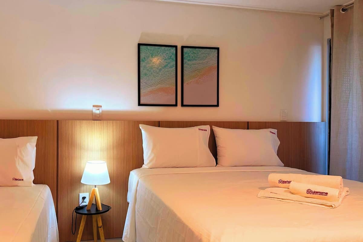 Quarto de hotel com duas camas, uma mesa de cabeceira com abajur e duas imagens emolduradas na parede.