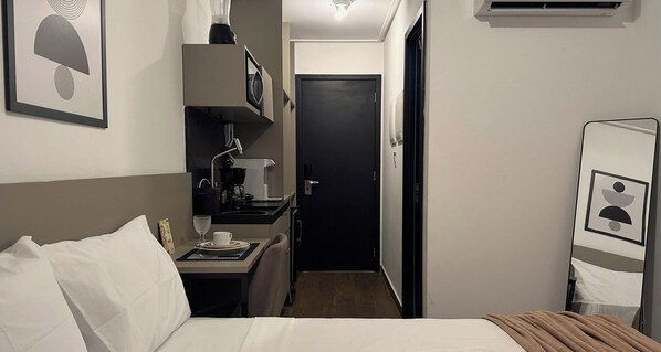 Quarto de hotel pequeno, com cama, escrivaninha com cafeteira, espelho e porta que dá para outro cômodo.
