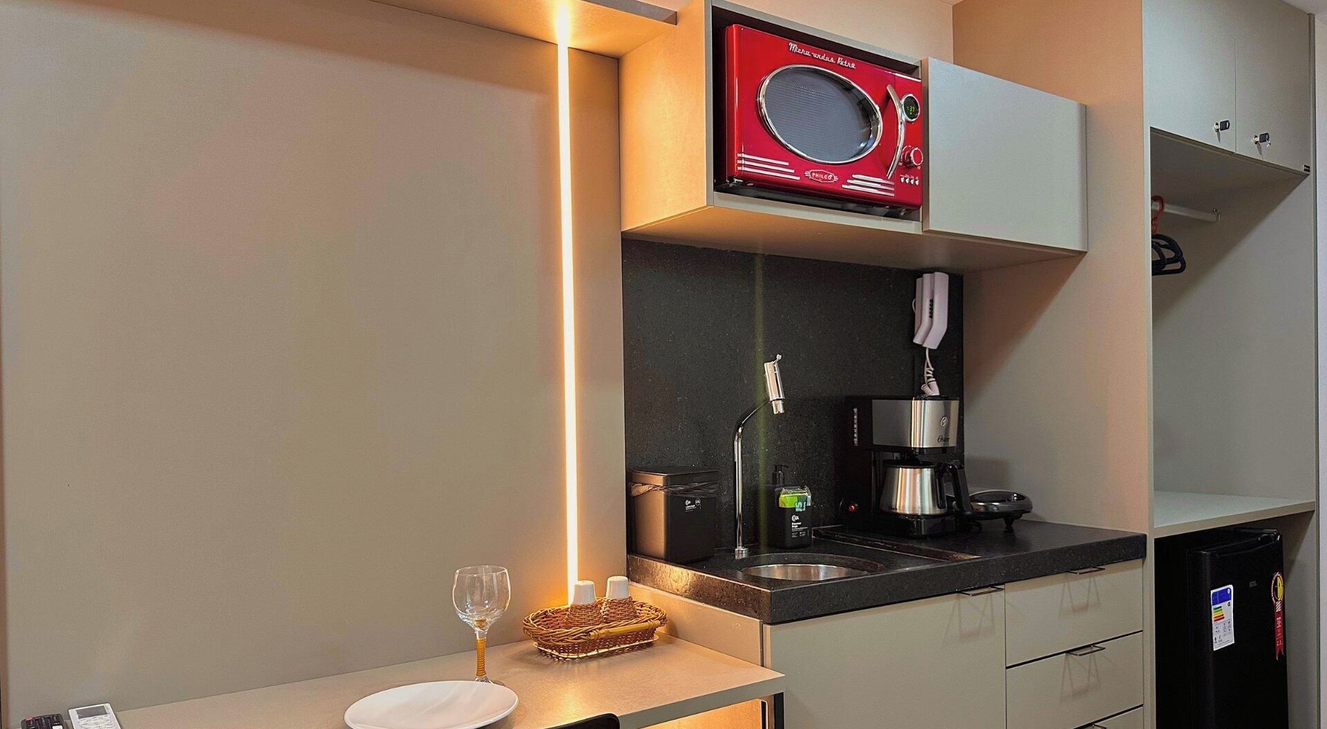 Cozinha compacta com micro-ondas, cafeteira e uma taça de vinho no balcão.