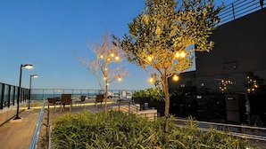 Een terras met stoelen en tafels, een boom met verlichting en uitzicht op de zee.