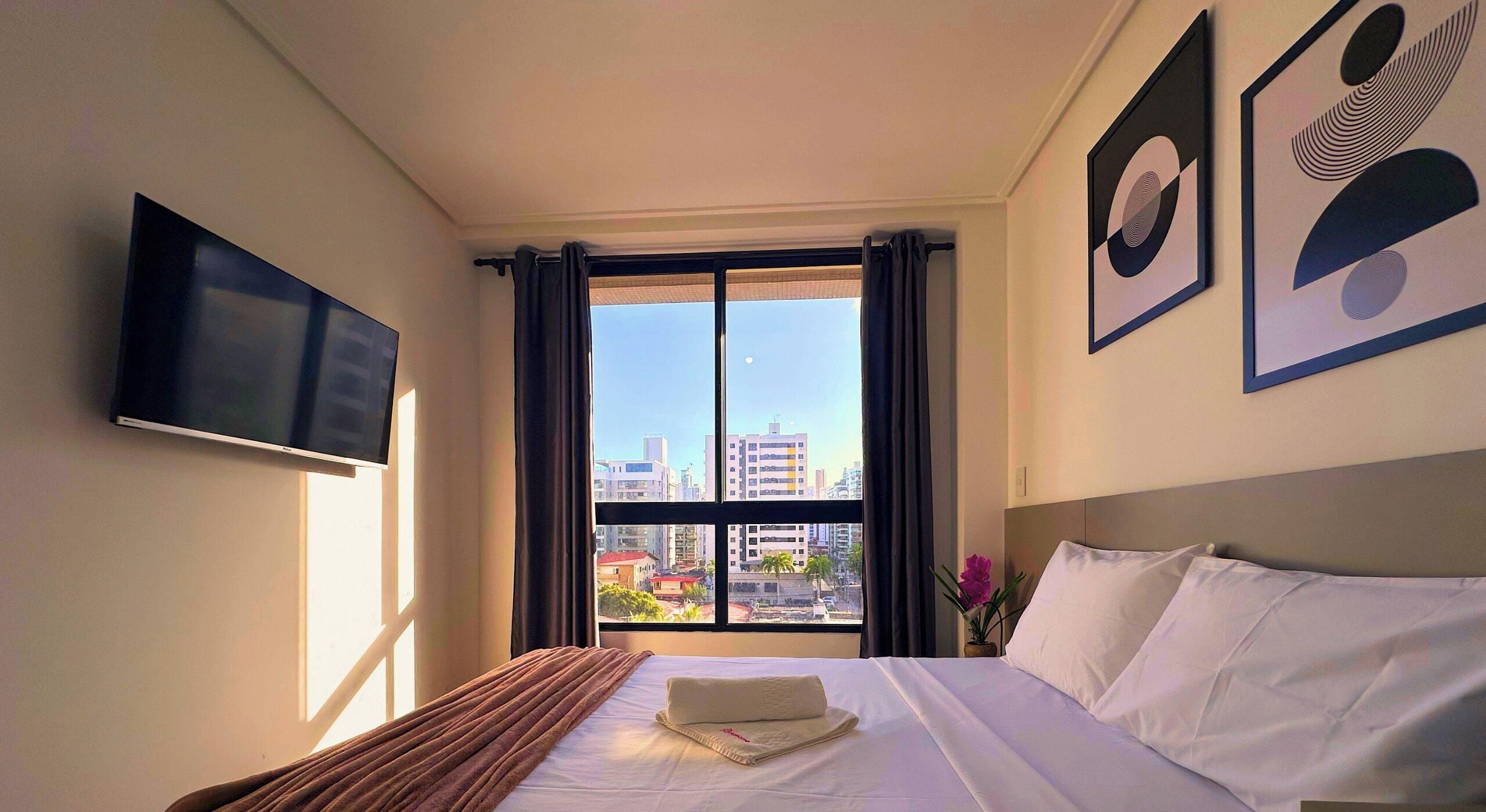 A hotel room with a bed, a flat-screen TV, a window with curtains, and artwork on the wall.
