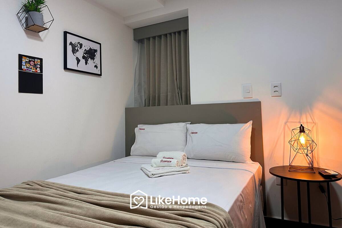 Una cama bien hecha con ropa de cama blanca y una toalla doblada, un mapa mundial en la pared, una pequeña planta en maceta y una lámpara en una mesita de noche.