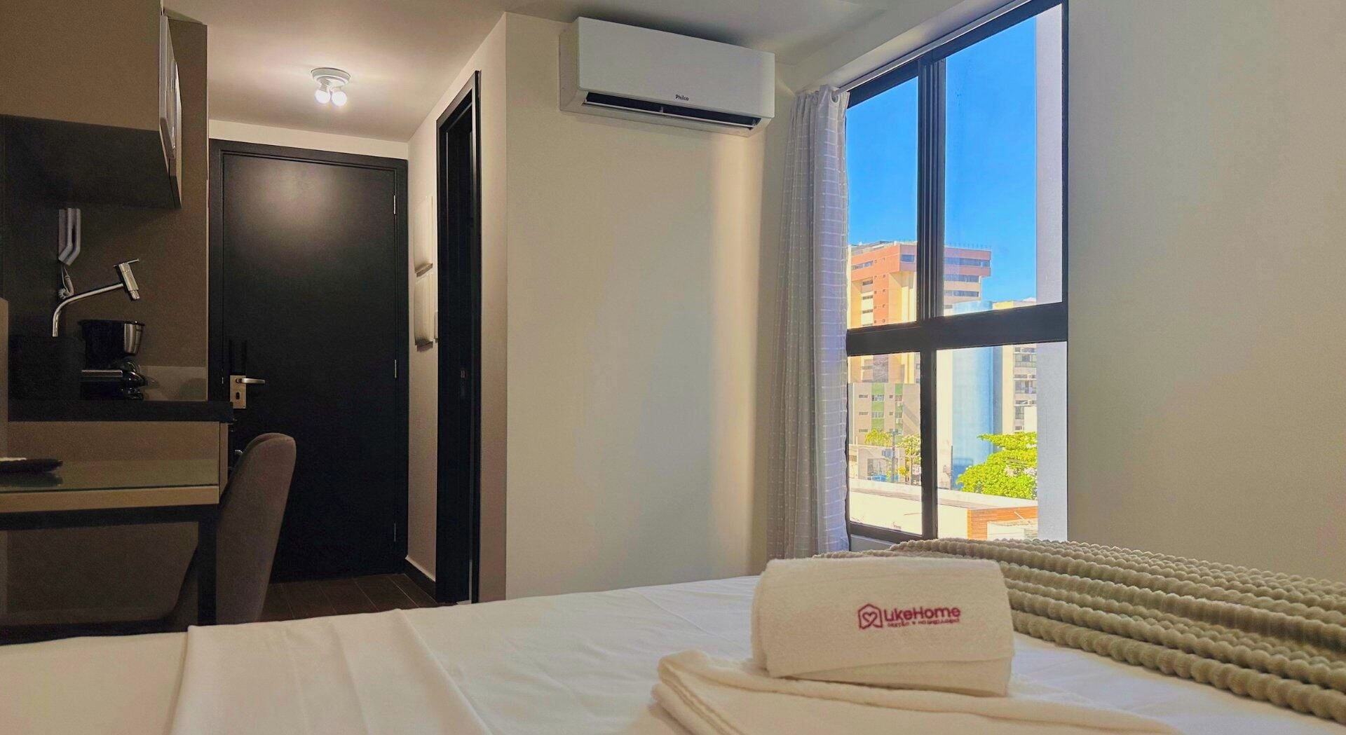 A hotel room with a bed, a desk, a chair, an air conditioning unit, and a window with a view of buildings.