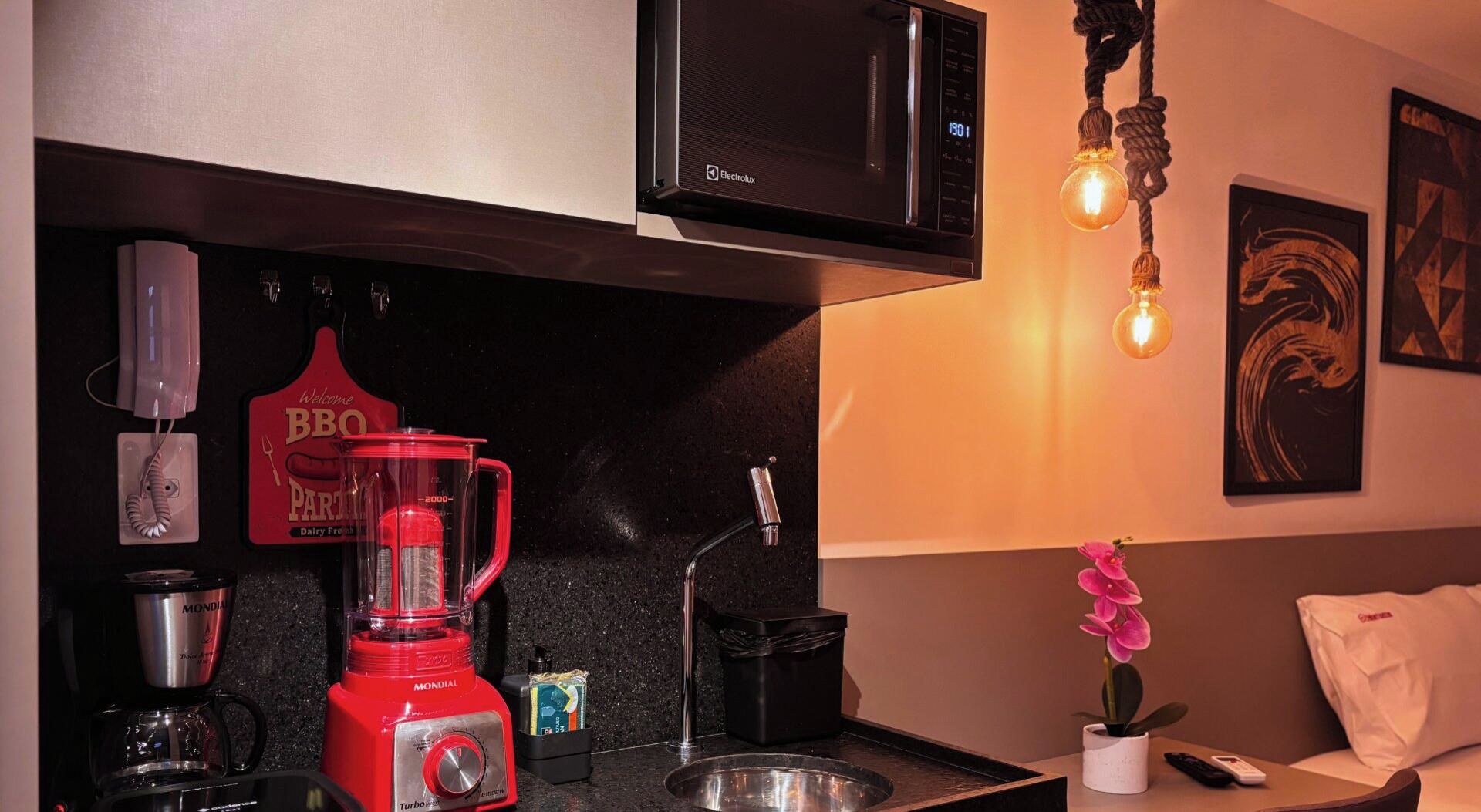 A kitchenette with a red blender, a coffee maker, a microwave, and a hanging light bulb.