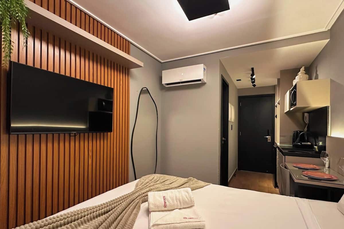 Habitación de hotel con cama, televisor montado en la pared, equipo de aire acondicionado, kitchenette pequeño y puerta que da a otra habitación.